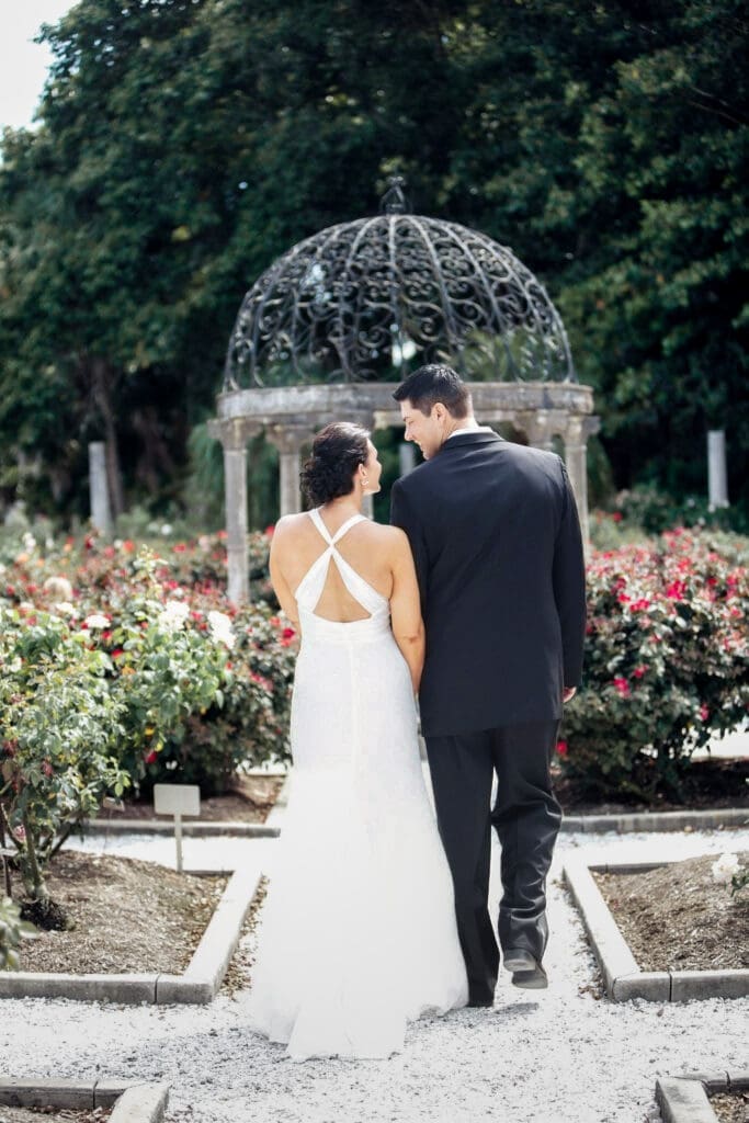 Bride and groom walking away together in The Ringling Museum gardens toward a romantic gazebo surrounded by roses